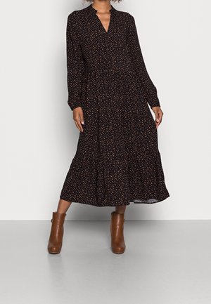 Kobieta ubrana w czarną midi sukienkę w małe pomarańczowe kropki z długimi rękawami, do której dobrała brązowe botki za kostkę, stojąca na tle jednolitego tła.