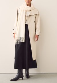 Beige trench coat with large collar and double-breasted buttons, layered over a white top. Black maxi skirt and fitted black ankle boots.