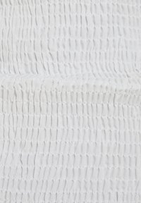 Bílá látka s texturovaným, nařaseným vzorem a rovnoměrně rozmístěnými záhyby, vytvářející jemný, splývavý vzhled.