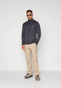 Dark gray zip-up jacket, beige cargo pants, and brown hiking boots. Accessories include white sunglasses. Smooth fabric and tailored fit.
