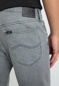Jeans de mezclilla gris con un acabado texturizado, que presentan una etiqueta de cuero negra en la cintura y bolsillos traseros cosidos, mostrando un diseño curvado.