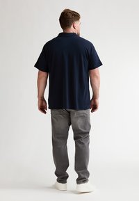 Polo bleu marine à manches courtes, en coton, associé à un jean gris. La personne porte des baskets blanches, vue de dos.