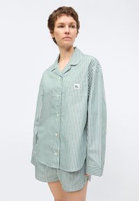 Γυναίκα με κοντά μαλλιά που φοράει πουκάμισο με κατακόρυφες ρίγες σε πράσινο και άσπρο και ταιριαστό σορτς, στέκεται μπροστά σε μονόχρωμο φόντο.