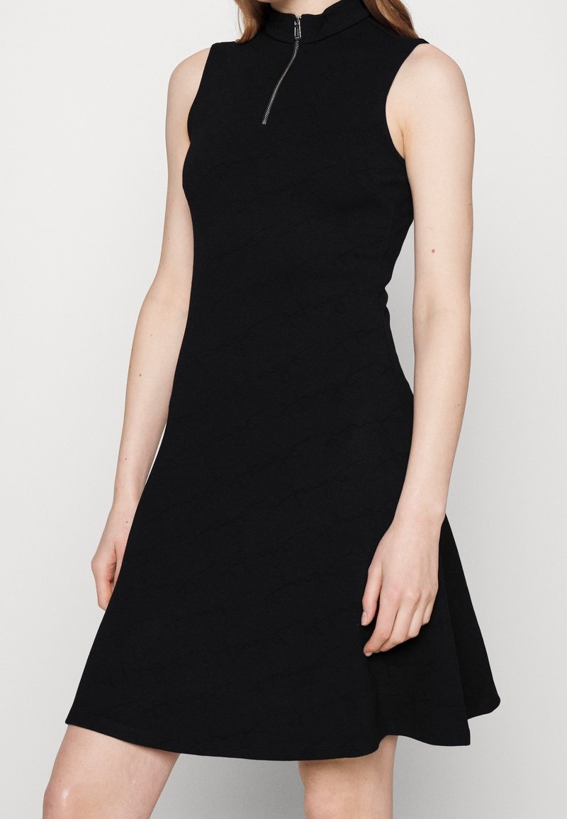 Robe noire sans manches avec un col montant et une fermeture éclair à l'avant, présentant un corsage ajusté et une jupe évasée, fabriquée dans un tissu lisse avec un motif subtil.
