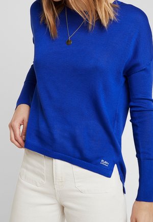 Femme portant un pull à manches longues bleu vif avec un petit logo blanc près de l'ourlet et un pantalon crème taille haute, avec un collier visible.