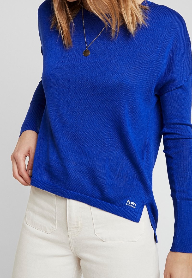 Femme portant un pull à manches longues bleu vif avec un petit logo blanc près de l'ourlet et un pantalon crème taille haute, avec un collier visible.