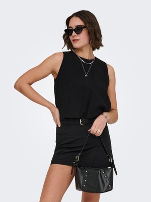 Woman in sleeveless black top and skirt wearing cat-eye sunglasses, layered necklaces, bracelet, ring, and holding a black studded handbag.