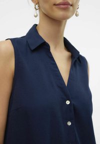 Mörkblå ärmlös blus med krage, framknäppning med vita knappar och slät textur.
