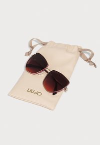 Sunglasses with large dark lenses, gold accents, and a burgundy frame, resting on a beige drawstring pouch with "LIU JO" printed in gold.