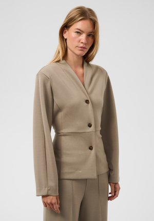 Femme aux cheveux blonds lisses portant une veste beige boutonnée et un pantalon assorti, debout devant un fond clair uni.