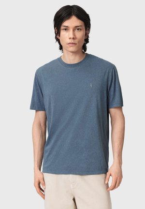 Nuori mies, jolla on suora tumma tukka, pukeutuu yksiväriseen siniseen lyhythihaiseen t-paitaan ja vaaleanbeigeihin housuihin yksinkertaista vaaleaa taustaa vasten.