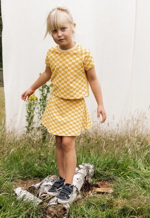 Geel geruite tweedelige outfit, bestaande uit een korte mouwen top en een uitlopende rok, gecombineerd met zwarte sneakers.