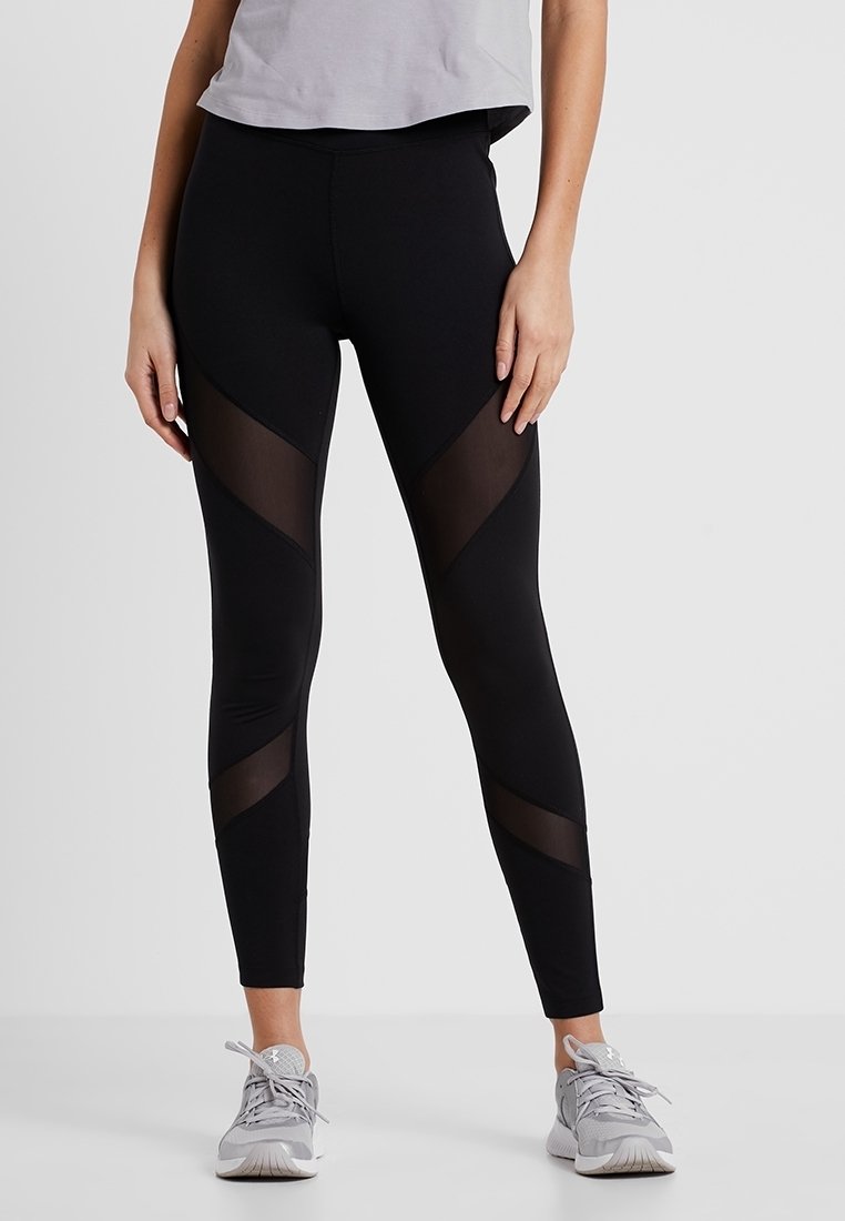 Svarta träningstights med genomskinliga mesh-paneler i zigzag-mönster, hög midja och en åtsittande design, kombinerat med grå sneakers.