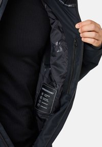 Zwart waterdichte jasinterieur met een gladde textuur, voorzien van een label dat de waterdichte, ademende en winddichte eigenschappen en getapete naden beschrijft.