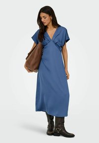Niebieska sukienka do kolan z krótkimi rękawami, głębokim dekoltem w kształcie litery V oraz fazowaną przednią częścią; zestawiona z brązową torbą na ramię i czarnymi botkami sięgającymi do kolan.