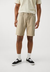 Shorts de mezclilla beige con un corte más relajado, que presentan bolsillos frontales y cierre de botón. Combinados con zapatillas negras y calcetines blancos hasta el tobillo.