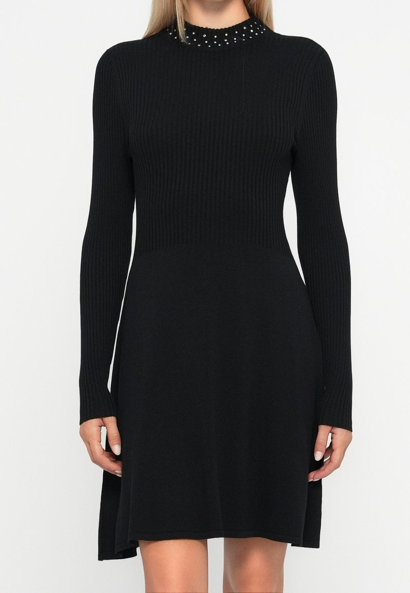 Femme portant une robe en maille noire à manches longues avec un haut côtelé ajusté et un détail de col perlé, debout sur un fond uni.