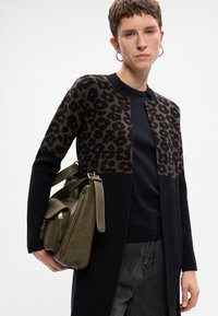 Osoba w czarno-brązowym kardiganie w print w panterkę, czarnej bluzce, ciemnych dżinsach, złotych kolczykach w kształcie kółek, trzymająca oliwkową skórzaną torebkę.