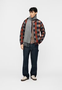 Νέος άνδρας που φοράει πολύχρωμο πουλόβερ καρό, ριγέ πουκάμισο, σκοτεινό τζιν, λευκά αθλητικά παπούτσια και γκρι κασκόλ, όρθιος με τα χέρια στις τσέπες.