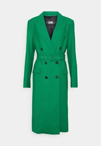 Groene dubbelzijdige trenchcoat met een ceintuur in de taille, zwarte knopen, inkepingen op de revers en twee voorzakken. Glad, gestructureerd materiaal.