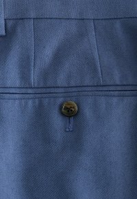 Couture en tissu bleu texturé avec un seul bouton sombre à quatre trous maintenu par un fil bleu sur un vêtement.