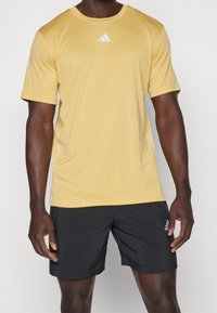 Muž v žlutém tričku s krátkým rukávem Adidas a černých šortkách Adidas, stojící před jednobarevným pozadím.