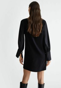 Crna saten haljina s dugim, napufanim rukavima i ravnim krojem. Ima prorez na leđima i dugmad kao ukras na manžetama, stvarajući elegantnu siluetu.