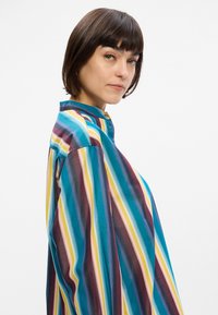 Πολύχρωμο πουκάμισο με ριγέ σχέδιο και ψηλό γιακά, με κάθετες γραμμές σε τιρκουάζ, μωβ, κίτρινο και ροζ. Ελαφρύ ύφασμα και χαλαρή εφαρμογή.