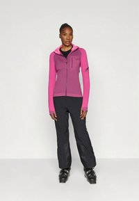 Sisehäälestatud lukuga jakk kahes roosa varjus, millel on kapuuts ja pikad varrukad, kombineeritud mustade suusapükste ning mustade suusasaabastega.