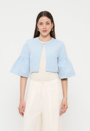 Žena nosící světle modrou krátkou bundu se širokými řasenými rukávy přes bílý top a vysokým pasem krémové kalhoty, stojící na bílém pozadí.