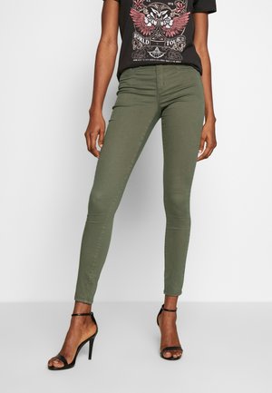 Person trägt olivgrüne Skinny-Hose, schwarzes T-Shirt mit Grafik und schwarze elegante Sandalen mit Knöchelriemen auf weißem Hintergrund.