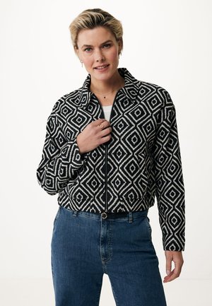Schwarz-weiße Zip-Jacke mit Rautenmuster, langen Ärmeln und Kragen; getragen über einem weißen Oberteil und kombiniert mit blauen Jeans.