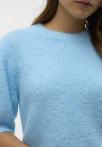 Gai zils adīts džemperis ar apaļu kakla izgriezumu, īsām piedurknēm un mīkstu, pūkainu tekstūru. Tam ir ribota apdare apkaklei un piedurknēm.