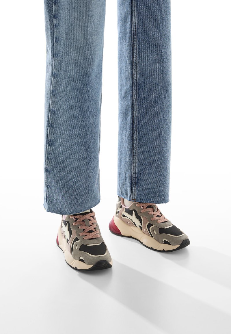 Persona che indossa jeans blu e sneakers multicolori beige, nere e rosa, in piedi su un pavimento bianco con ombre.