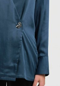 Blusa de satén azul con un tacto suave, presenta un destacado botón plateado en forma de corazón y mangas largas con puños anchos.