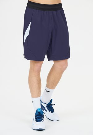 Ben iført marineblå atletiske shorts med sort taljebånd, hvide mellemlange strømper og hvide og blå løbesko, stående på hvid baggrund.