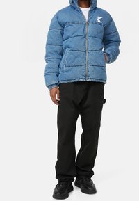 Blue denim puffer jacket with a hood, zip closure, ribbed cuffs, and logo detail. Worn over black pants and black sneakers.