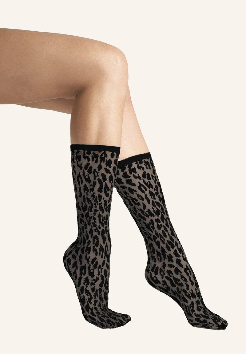 Ben klædt i gennemsigtige sorte netstrømper med mørkt leopardmønster, vist mod en enkel lys baggrund.