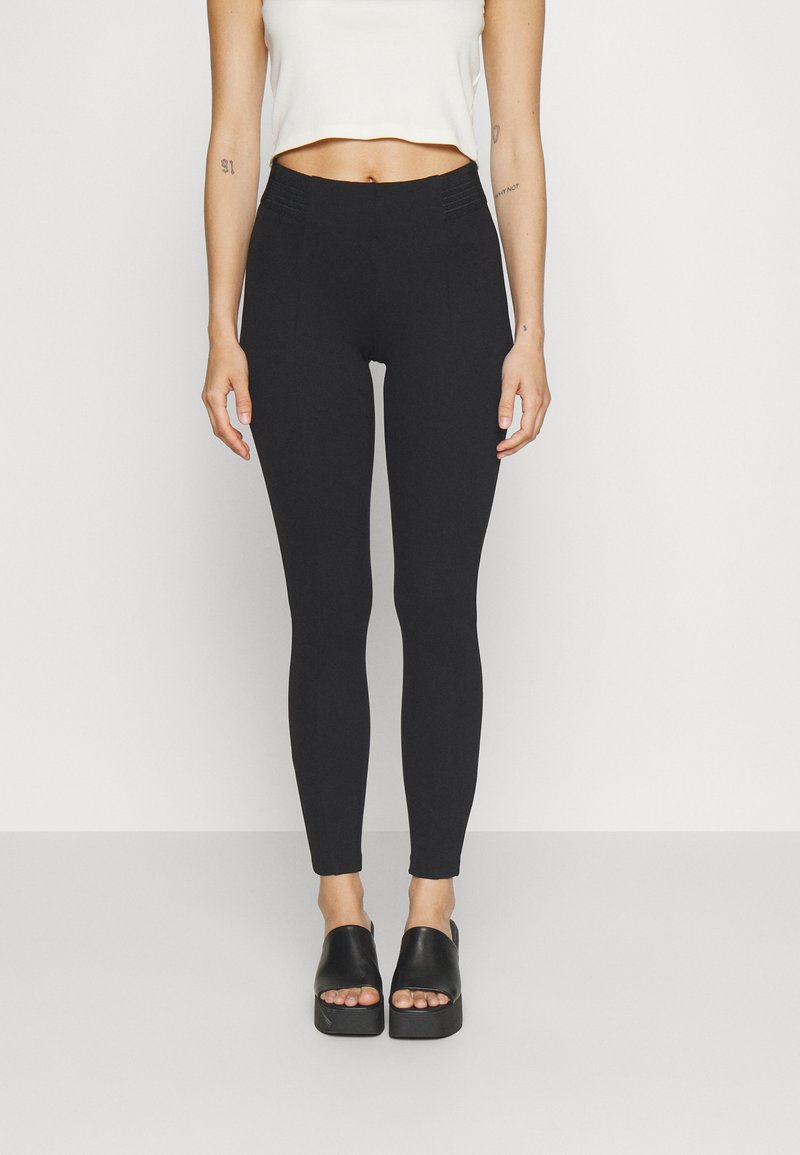 Fekete, magas derekú leggings, rugalmas anyagból, sima textúrával és szoros illeszkedéssel, fekete, vastag talpú csúszó szandálokkal párosítva.