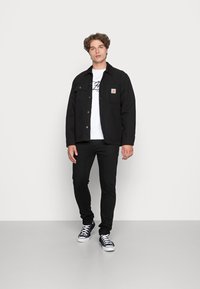 Zwarte jas met knoopsluitingen en voorzakken, gecombineerd met een wit t-shirt en skinny zwarte jeans; model draagt zwarte sneakers.