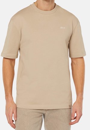 Mies, joka käyttää beigeä lyhythihasta pyöreäkauluksista T-paitaa, jossa vasemmassa rinnassa pieni valkoinen "B939"-logo, yhdistettynä vaalean beigen värisiin housuihin.