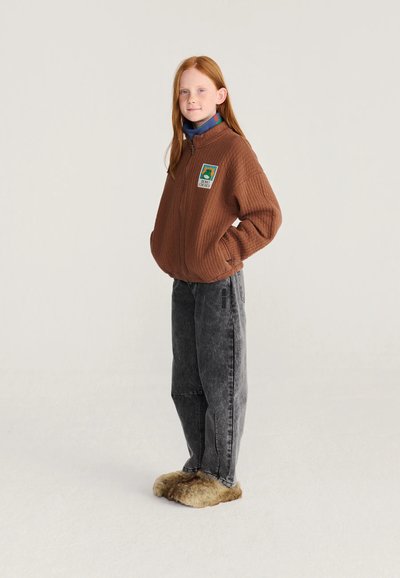 Jeune fille aux cheveux roux portant une veste marron zippée, un jean large gris foncé et des chaussons duveteux, debout les mains dans les poches.