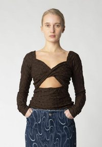 Brun langærmet bluse med tekstureret stof, der har et nøglehuldesign og en tætsiddende silhuet. Båret med en højtaljet denimnederdel med mønster i blå.