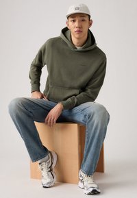 Φούτερ με κουκούλα σε ελιά πράσινο χρώμα με εμπρός τσέπη, συνδυασμένο με ανοιχτό μπλε τζιν και γκρι αθλητικά παπούτσια, καθισμένος σε ξύλινο κάθισμα. Ουδέτερο φόντο.