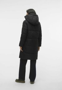 Manteau noir matelassé avec capuche, présentant un design mi-long, une texture lisse et des coutures horizontales. Porté avec un jeans large noir.