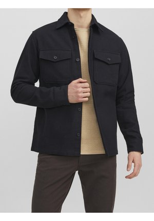 Mann trägt schwarze, geknöpfte Jacke mit zwei Brusttaschen über beigem, strukturiertem Pullover und dunklen Hosen, Hand hält die Vorderseite der Jacke.