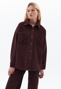 Burgundy manchester skjorta med krage, två framfickor och knäppningar; har en avslappnad passform och ribbad textur.