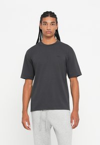 Νεαρός άνδρας με πλεξούδες, φορώντας ένα σκούρο γκρι oversized T-shirt και ανοιχτό γκρι παντελόνι φόρμας, στέκεται μπροστά σε ένα λευκό φόντο.