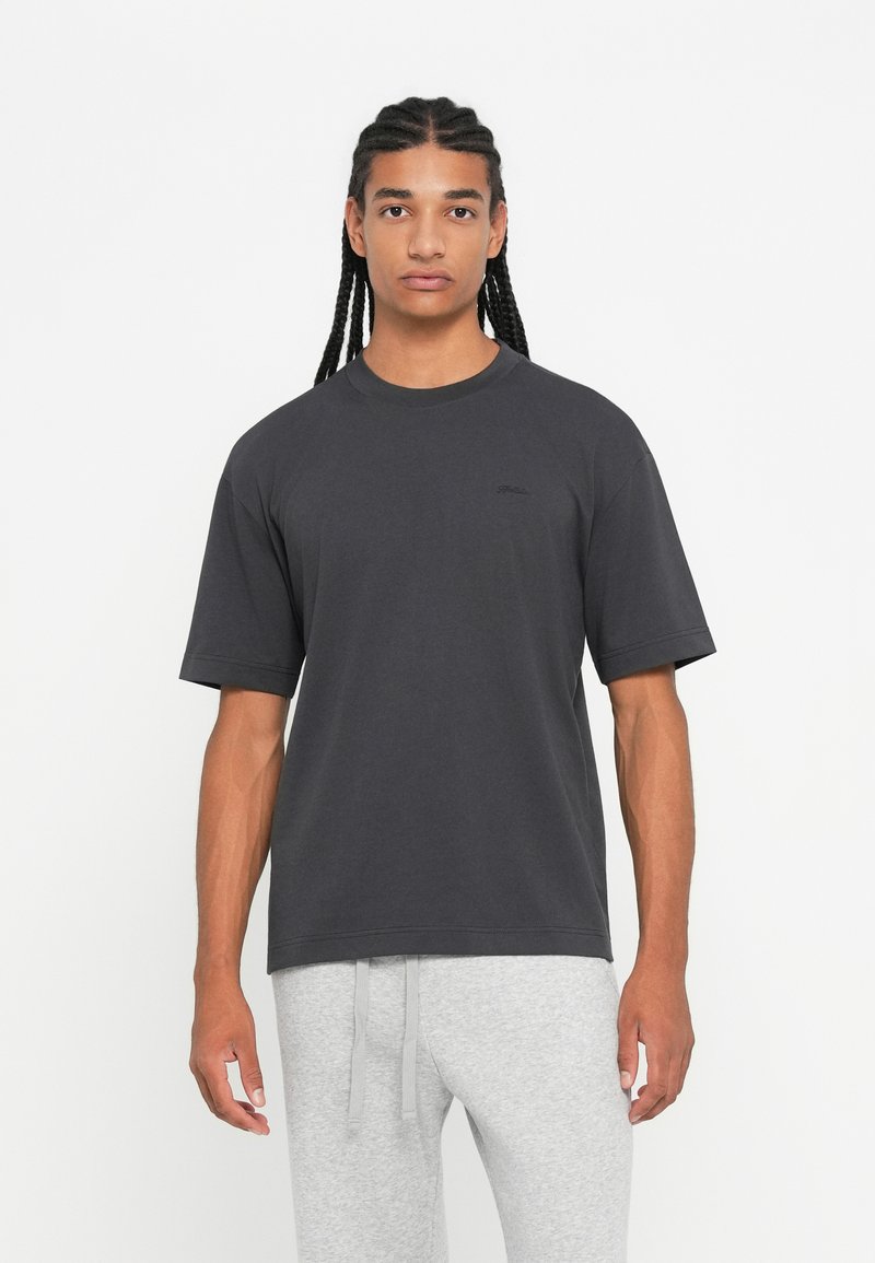 Νεαρός άνδρας με πλεξούδες, φορώντας ένα σκούρο γκρι oversized T-shirt και ανοιχτό γκρι παντελόνι φόρμας, στέκεται μπροστά σε ένα λευκό φόντο.