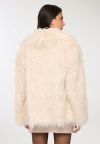 Cappotto in finta pelliccia color crema con vestibilità ampia, grande cappuccio e maniche lunghe, caratterizzato da una consistenza soffice e un orlo grezzo.